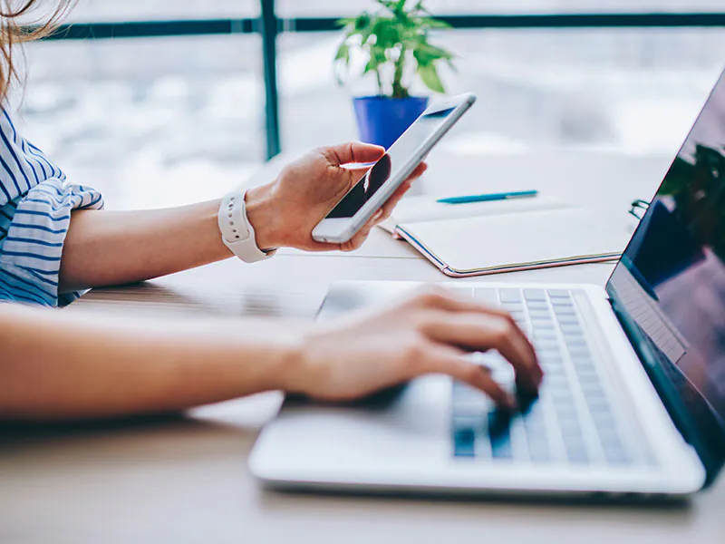 Woman is working on a laptop and is holding her smartphe in the left hand
